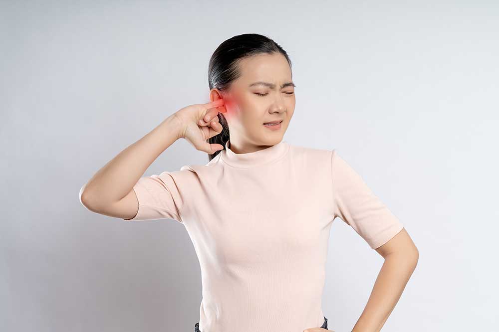Young woman in a pink top wincing as she presses a finger into her clogged ear, a concern Pacific Coast Audiology sees in San Luis Obispo, CA